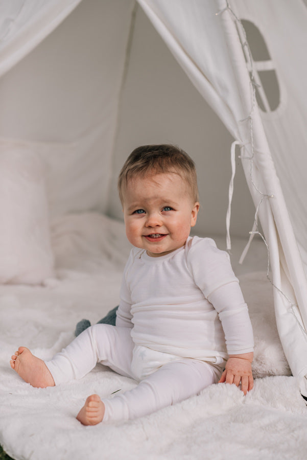 A baby sits in a white teepee looking at the camera and smiling.