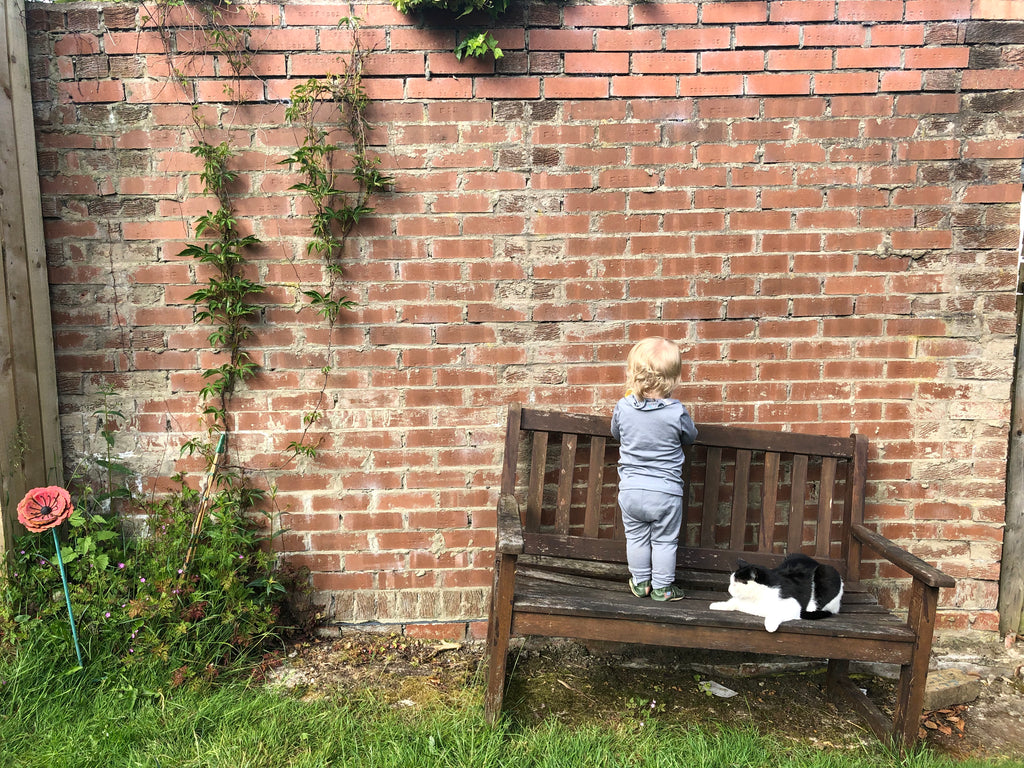 A baby stands on a garden bench facing the brick wall behind it