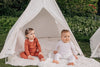 two babies sit on rugs outside a play teepee wearing wool pyjamas