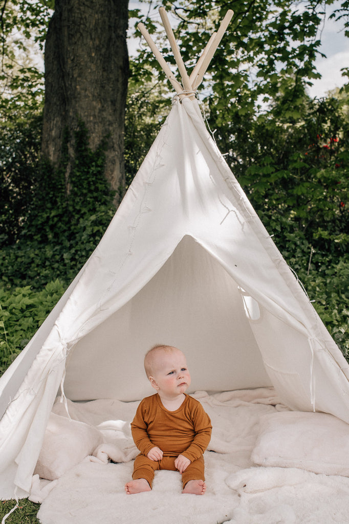 A baby sits in front of a play teepee outside, wearing a merino sleepsuit in ochre.
