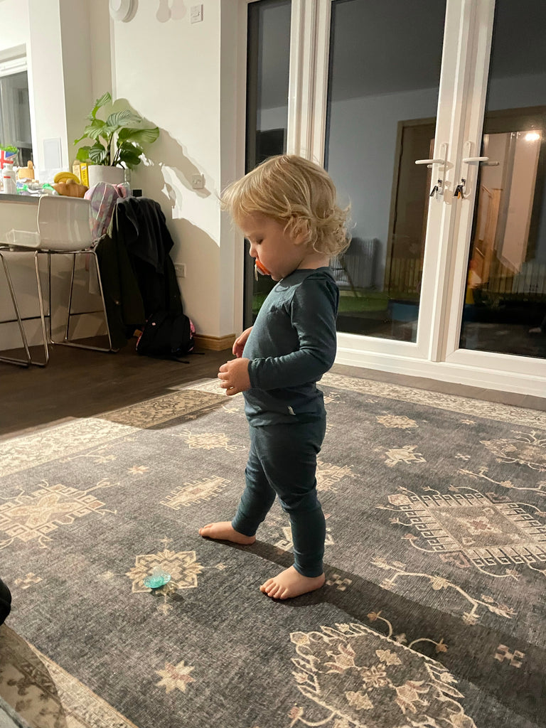 A toddler stands in a living room wearing soft teal merino wool pyjamas